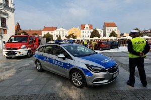 Policjant ruchu drogowego stojący na płycie chełmińskiego rynku podczas pokazów współdziałania służb ratowniczych. Widać również radiowóz oraz wóz strażacki.