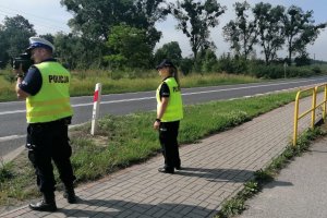 policjant oraz policjantka stoją przy drodze i obserwują ruch pojazdów. Policjant mierzy prędkość