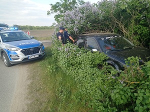 czarny pojazd stoi w zaroślach przy nim stoją umundurowani policjanci a na drodze ustawiony jest radiowóz