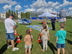 dzieci i mężczyzna stoją przed plansza do gry plenerowej z oddali patrzy na nich policjant